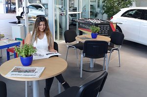 Wartebereich des Duderst&auml;dter Mercedes-Autohauses mit einer jungen Frau, die eine Zeitschrift liest, mehreren Tischen mit Zeitungen und Pflanzen, einem Kicker-Tisch im Hintergrund sowie einem wei&szlig;en Auto, das durch eine Glaswand sichtbar ist.