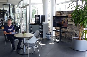 Heller Wartebereich im Mercedes-Autohaus mit Glasfront. Ein junger Mann sitzt an einem runden Tisch und liest eine Brosch&uuml;re. Neben ihm stehen weitere St&uuml;hle, ein kleiner Tisch mit einer Pflanze, ein Kaffeeautomat mit Tassenregal und ein gro&szlig;er Fernseher. Rechts im Bild eine gro&szlig;e gr&uuml;ne Pflanze in einem grauen Topf.