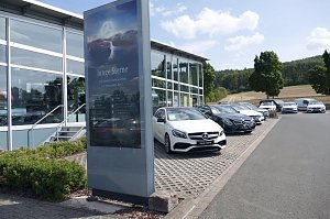 Reihe von Mercedes-Benz Gebrauchtwagen (Junge Sterne) auf dem Parkplatz eines Autohauses. Im Vordergrund steht ein gro&szlig;es Schild mit dem Logo und der Aufschrift �Junge Sterne � Die besten Gebrauchten von Mercedes-Benz�. Im Hintergrund moderne Glasfassade des Autohauses, B&auml;ume und Felder bei sonnigem Wetter.