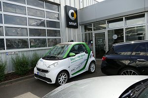 Weiß-grüner Smart mit der Aufschrift ‚Smart unter Strom und die Zukunft gehört DIR!‘, geparkt vor einem Autohaus. Auf dem Auto ist ein Blitzmotiv abgebildet. Im Hintergrund ist das Smart-Logo an der Gebäudewand zu sehen.
