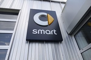 Nahaufnahme eines schwarzen Schildes mit dem Smart-Logo an einer silberfarbenen Metallfassade. Das Logo besteht aus einem stilisierten grauen ‚C‘ mit einem gelben, pfeilförmigen Element, darunter steht der Schriftzug ‚smart‘ in weißen Buchstaben.