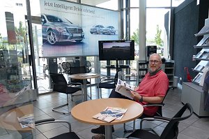 Heller Wartebereich in einem Mercedes-Benz-Autohaus. Ein Mann sitzt an einem runden Tisch, liest eine Brosch&uuml;re und schaut in die Kamera. Neben ihm liegen Magazine auf dem Tisch. Im Hintergrund ein gro&szlig;er Werbebanner mit Mercedes-Autos, ein Fernseher, St&uuml;hle, ein Kaffeeautomat und Regale mit Prospekten.