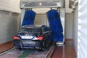 Ein schwarzer Mercedes-Benz wird in einer automatischen Waschanlage mit blauen B&uuml;rsten gereinigt. Das Fahrzeug ist mit Schaum bedeckt, die Anlage befindet sich in einer gefliesten Halle.