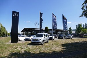 Mercedes-Benz Autohaus in Northeim mit gro&szlig;en Glasfronten, mehreren ausgestellten Fahrzeugen vorne, darunter wei&szlig;e Transporter und Smart-Modelle. Im Vordergrund hohe Mercedes-Flaggen und ein schwarzer Mercedes-Stern-Pylon. Dahinter der gro&szlig;e Mercedes-Stern auf dem Dach und eine gr&uuml;ne Heckenskulptur in Sternform. Blauer Himmel, sonniges Wetter.