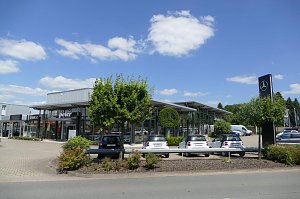 Autohaus mit gro&szlig;er Glasfassade und den Logos von Smart und Peter. Auf dem Vorplatz sind mehrere geparkte Smart-Autos zu sehen. Rechts steht ein hoher schwarzer Mast mit dem Mercedes-Benz-Stern. Vor dem Geb&auml;ude befinden sich B&auml;ume und B&uuml;sche. Der Himmel ist blau mit vereinzelten Wolken.