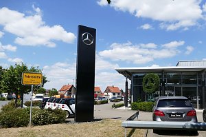 Mercedes-Autohaus mit gro&szlig;er Glasfassade und Mercedes-Stern aus Buschwerk. Links ein gelbes Ortsschild mit der Aufschrift �Petersh&uuml;tte Stadt Osterode am Harz�. Mehrere geparkte Autos, blauer Himmel mit wei&szlig;en Wolken.