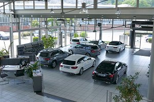 Gro&szlig;er, heller Showroom des Autohauses mit gro&szlig;en Glasfronten. Mehrere Mercedes-Benz Fahrzeuge in verschiedenen Farben (schwarz, silber, wei&szlig;) stehen ausgestellt. Links B&uuml;rotische mit Pflanzen und Unterlagen. Drau&szlig;en sind weitere Autos und Gr&uuml;nfl&auml;chen sichtbar. Die Atmosph&auml;re wirkt modern und einladend.