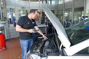 Zwei M&auml;nner stehen in einer Werkstatt neben einem Auto mit ge&ouml;ffneter Motorhaube. Der Mann links tr&auml;gt ein schwarzes Poloshirt mit "Peter" Logo und zeigt mit einem Tablet auf den Motor. Der andere Mann tr&auml;gt ein kariertes Hemd und schaut interessiert. Im Hintergrund sieht man Werkstatt-Ausr&uuml;stung, Werkb&auml;nke und Glasw&auml;nde mit Blick in weitere Bereiche der Werkstatt.