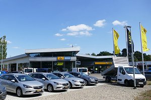 Au&szlig;enansicht des Opel-Autohauses in Osterode mit gro&szlig;em Vorplatz. Im Vordergrund stehen mehrere silberfarbene Opel-Pkw und ein wei&szlig;er Opel-Transporter mit Kipper-Aufbau. Vor dem Geb&auml;ude wehen gelbe und schwarze Opel-Flaggen. Das Autohaus hat eine moderne Glasfassade, dar&uuml;ber sind Schilder mit Opel Service und dem Opel-Logo sichtbar. Der Himmel ist blau mit einigen kleinen Wolken.