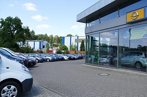 Au&szlig;enansicht des Opel-Autohauses mit Glasfassade. Rechts ist das gelbe Opel-Logo &uuml;ber dem Eingang sichtbar. Vor dem Geb&auml;ude sind viele Autos geparkt, darunter Transporter und verschiedene Pkw-Modelle in einer Reihe. Im Hintergrund sieht man B&auml;ume und Industriegeb&auml;ude. Der Himmel ist blau mit einigen Wolken.
