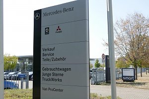 Gro&szlig;es Schild vor dem Mercedes-Benz Autohaus Peter in Dessau. Auf dem Schild stehen die Bereiche: Verkauf, Service, Teile/Zubeh&ouml;r, Gebrauchtwagen, Junge Sterne, TruckWorks und Van ProCenter. Oben Logos von Mercedes-Benz, Smart und Fuso. Im Hintergrund Glasfassade des Autohauses, parkende Autos und ein Baum.