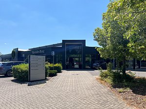 Der moderne Eingangsbereich des Autohauses mit großen Fenstern und der Aufschrift Mercedes-Benz. Im Vordergrund steht ein Wegweiser mit den Bereichen: Verkauf, Service und Teile/Zubehör. Rechts steht ein Baum mit grünen Blättern.
