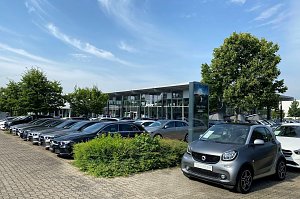 Au&szlig;enansicht des Mercedes-Benz Autohauses Peter in Dessau. Im Vordergrund steht ein grauer Smart mit dem Schild �Gebrauchtwagen�. Daneben viele schwarze und silberne Mercedes-Benz Fahrzeuge in einer Reihe auf dem Parkplatz. Im Hintergrund modernes Glasgeb&auml;ude mit gro&szlig;en Fenstern, B&auml;umen und blauem Himmel mit leichten Wolken.