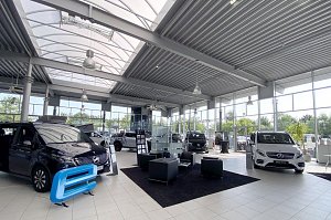 Gro&szlig;er, heller Showroom eines Mercedes-Benz Autohauses mit hohen Glasfronten und modernen Deckenleuchten. Mehrere Vans und Transporter sind ausgestellt, darunter ein wei&szlig;er und ein schwarzer Mercedes. In der Mitte ein schwarzer Teppich mit vier schwarzen Sesseln und einem Tisch als Wartebereich. Viel nat&uuml;rliches Licht durch die gro&szlig;en Fenster.