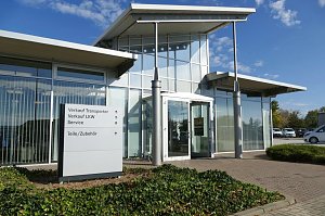 Glasfassade des Mercedes-Autohauses in Zerbst mit modernem Eingangsbereich, daneben ein Wegweiser mit Hinweisen zu Verkauf, Service und Zubeh&ouml;r, davor niedrige Bodendecker-Pflanzen, blauer Himmel im Hintergrund.