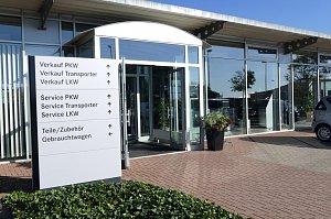 Eingangsbereich des Mercedes-Benz Autohauses in Bernburg mit gro&szlig;en Glasfronten und offen stehender Glast&uuml;r, davor ein wei&szlig;es Hinweisschild mit Richtungsangaben zu Verkauf, Service, Zubeh&ouml;r und Gebrauchtwagen, daneben Blumenk&uuml;bel, gepflasterter Boden und blauer Himmel.