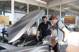 Ein Mitarbeiter im schwarzen Hemd mit Mercedes-Logo erkl&auml;rt einem jungen Kunden Details am Motorraum eines grauen Mercedes-Fahrzeugs.