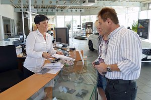 Mitarbeiterin am Empfangstresen im Mercedes-Benz Autohaus Bernburg &uuml;bergibt l&auml;chelnd Unterlagen an ein Paar, moderner heller Showroom mit Computern, Prospekten und Autos im Hintergrund.