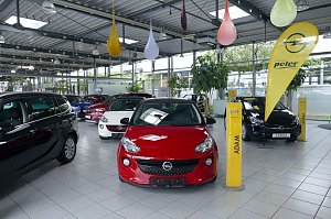 Innenansicht eines Autohauses mit mehreren ausgestellten Opel-Fahrzeugen, darunter ein roter Opel Adam im Vordergrund und ein schwarzer Opel Corsa im Hintergrund; gelbe Opel-Werbeflaggen und bunte Dekoelemente hängen von der Decke.