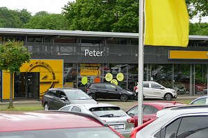 Ansicht des Opel Autohauses Peter mit großen Glasfassaden, gelbem Opel-Logo über dem Eingang und mehreren geparkten Autos im Vordergrund; gelbe Fahne und Bäume im Hintergrund.