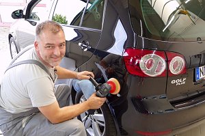 Pierre Heyer poliert die Heckseite eines schwarzen VW Golf Plus. Er trägt graue Arbeitskleidung, hält eine Poliermaschine in der Hand und schaut lächelnd in die Kamera. Das Fahrzeug glänzt stark, Spiegelungen sind deutlich zu sehen.