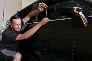 Kurt Kuschmierz arbeitet lächelnd an der Tür eines schwarzen Autos. Er benutzt ein Spezialwerkzeug mit gelbem Griff, um eine Delle vorsichtig auszubeulen. Er kniet neben dem Fahrzeug und zeigt dabei Feingefühl und Präzision. Werkstattumgebung im Hintergrund.