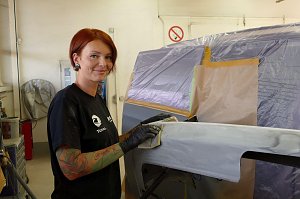 Katja Röpcke führt Schleifarbeiten an einem grundierten Karosserieteil durch. Sie trägt schwarze Handschuhe und ein schwarzes T-Shirt mit Firmenlogo, hat rote Haare und sichtbare Tattoos am Arm. Im Hintergrund ein abgeklebtes Fahrzeug in einer Werkstatt.