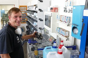 Peter Meyer lächelt, während er an einer Farbmischstation arbeitet. Er hält eine Lackierpistole und trägt eine Atemschutzmaske um den Hals. Vor ihm ein Monitor, viele Farbdosen und große Kanister. Im Hintergrund Regale mit weiteren Farben und Materialien.