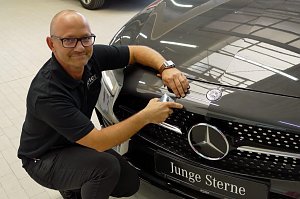 Dirk Morgenthal, Leiter des Lackierzentrums, kniet neben einem schwarzen Mercedes mit dem Schild ‚Junge Sterne‘. Er trägt ein schwarzes Polohemd mit Aufschrift ‚Peter‘ und und hält ein Farbmessgerät an die Motorhaube, um den exakten Farbton zu bestimmen. Heller Werkstattboden, reflektierende Autolackierung.