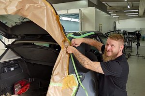 Frank Frank lächelt, während er ein Fahrzeug für Lackierarbeiten mit braunem Abdeckpapier abklebt. Das Auto steht mit geöffneter Heckklappe in einer Werkstatt. Im Hintergrund sind weitere Fahrzeuge und Arbeitsbereiche zu sehen.