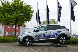 Silberner Peugeot 3008 mit Werbebeschriftung auf einer erh&ouml;hten Pr&auml;sentationsrampe vor einem Autohaus. Im Hintergrund wehen mehrere Peugeot-Flaggen und es steht ein Baum sowie ein Geb&auml;ude mit roten und grauen Fassadenelementen.