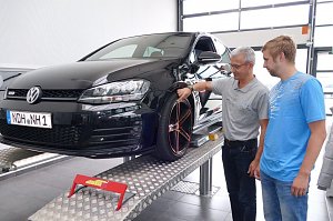 Schwarzer VW Golf GTD steht auf einer Hebeb&uuml;hne in einer Werkstatt. Ein Mitarbeiter in grauem Poloshirt zeigt einem Kunden in blauem T-Shirt Details am Rad. Beide l&auml;cheln und wirken im Gespr&auml;ch vertieft.