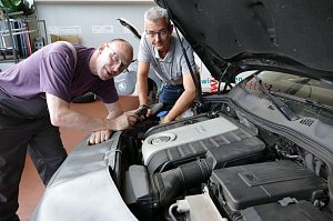 Zwei Mitarbeiter der Autowelt Peter GmbH arbeiten gemeinsam an einem VW-Motor. Einer h&auml;lt ein Diagnoseger&auml;t, der andere beugt sich &uuml;ber den Motorraum. Beide tragen Arbeitskleidung und wirken konzentriert.