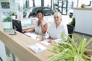 Zwei Serviceassistentinnen sitzen am Empfangstresen in einem modernen Autohaus. Eine Frau telefoniert und l&auml;chelt, die andere schreibt etwas auf. Beide tragen wei&szlig;e Blusen. Auf dem Schreibtisch stehen ein Monitor, Unterlagen sowie Pflanzen. Im Hintergrund sind Autos und der helle Ausstellungsraum zu sehen.