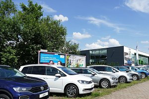 Reihe von VW-Fahrzeugen in unterschiedlichen Farben, die auf einem Parkplatz vor einem modernen Autohaus mit Glasfassade stehen. Im Hintergrund sind Werbetafeln und gr&uuml;ne B&auml;ume zu sehen, der Himmel ist blau mit einigen Wolken.