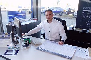 Autohaus-Mitarbeiter in wei&szlig;em Hemd sitzt an einem Schreibtisch vor einem Computer, l&auml;chelt in die Kamera, Verkaufsunterlagen und Brosch&uuml;ren auf dem Tisch, Autos sind drau&szlig;en durch das Fenster sichtbar.