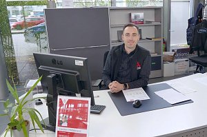 Mitarbeiter im Autohaus sitzt lächelnd an einem Schreibtisch mit Computer, Unterlagen und Pflanze, im Hintergrund Regale und Fenster mit Blick nach draußen.