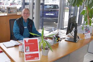 Ein Mitarbeiter sitzt freundlich lächelnd an einem Schreibtisch im Autohaus Peter in Göttingen, mit Namensschild und Pflanzen auf dem Tisch, im Hintergrund ein Auto mit Peter-Nummernschild.