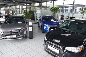 Innenansicht eines Autohauses mit mehreren Mitsubishi-Fahrzeugen in verschiedenen Farben, darunter ein grauer, ein blauer und ein schwarzer Wagen, auf glänzendem Fliesenboden mit großen Pflanzen und Glaswänden im Hintergrund.