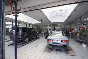Helle Mercedes-Benz Werkstatt mit zwei Fahrzeugen auf Hebeb&uuml;hnen: links ein schwarzer Gel&auml;ndewagen (G-Klasse), rechts ein silberner Oldtimer. Im Hintergrund Glasb&uuml;ros und Regale mit Autopflegeprodukten.