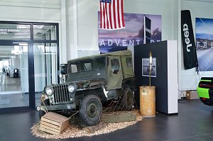 Historischer Jeep Willy in nostalgischer Präsentation im Showroom mit US-Flagge und Jeep-Banner im Hintergrund.