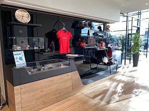 Mercedes-Benz Collection: Verkaufsbereich im Autohaus mit Verkaufsregalen, auf denen Mercedes-Fanartikel wie Shirts, Uhren und M&uuml;tzen ausgestellt sind. Viel Tageslicht durch gro&szlig;e Fenster.