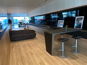 Moderner Kundenwartebereich im Autohaus mit Holzfu&szlig;boden, dunkelbraunem Sofa, Hochtisch mit Barhockern, historischen Fotos und Blick auf ein klassisches Auto im Hintergrund.