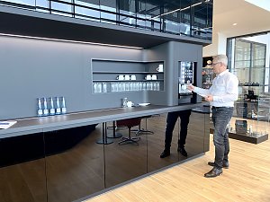 Mann im wei&szlig;en Hemd zapft Kaffee am modernen Kaffeeautomaten in einer stilvollen Autohaus-Lounge mit Gl&auml;sern, Tassen und Wasserflaschen.