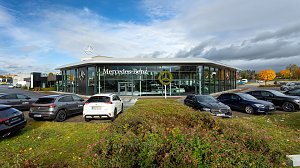 Mercedes-Benz Autohaus mit moderner Glasfassade und gro&szlig;em Mercedes-Logo, mehrere geparkte Fahrzeuge im Vordergrund, herbstliche B&auml;ume und Wolkenhimmel im Hintergrund.