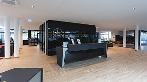 Moderner Empfangsbereich eines Mercedes-Benz Autohauses mit heller Holzoptik, schwarzem Empfangstresen, Glastrennw&auml;nden und Mercedes-Logo an der Wand.