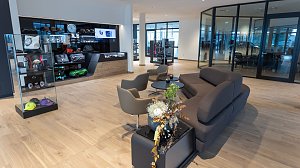 Moderner Wartebereich in einem Mercedes-Benz Autohaus mit bequemer grauer Couch, Sesseln, Glastischen, Blumenarrangement und Vitrine mit Mercedes-Accessoires und Fanartikeln.