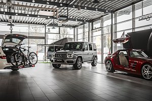 Innenraum einer modernen Mercedes-Benz-Ausstellungshalle mit gro&szlig;en Glasfronten. Zu sehen sind ein wei&szlig;er Kombi mit offenem Kofferraum und Fahrradtr&auml;ger, ein silberner Mercedes G-Klasse SUV und ein roter Sportwagen mit ge&ouml;ffneten Fl&uuml;gelt&uuml;ren. Heller, industrieller Boden und Decke mit Lampen.