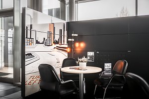 Gem&uuml;tliche Sitzecke mit rundem Tisch und vier schwarzen Lederst&uuml;hlen in einem Autohaus. Auf dem Tisch steht eine kleine Pflanze. Im Hintergrund eine Wand mit gro&szlig;em Stadtmotiv in Wei&szlig;- und Oranget&ouml;nen. Helle Fensterfront und moderner schwarzer Wandbereich.