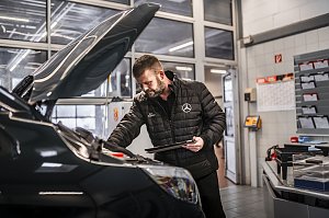 Werkstattmitarbeiter im Mercedes-Benz Autohaus pr&uuml;ft den Motorraum eines Fahrzeugs mit ge&ouml;ffnetem Vorderhaube. Er tr&auml;gt eine schwarze Jacke mit Mercedes-Logo und h&auml;lt ein Tablet. Im Hintergrund moderne Werkstatteinrichtung mit Dokumentenf&auml;chern, Kalender und Glast&uuml;ren.